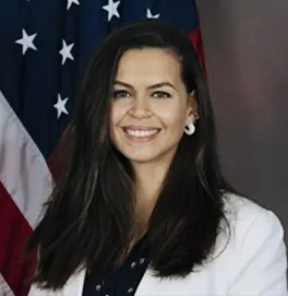 a woman in a white blazer smiles in front of an american flag