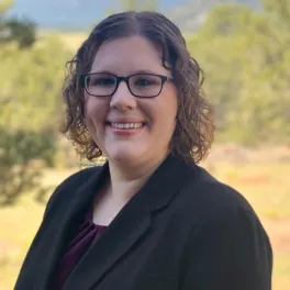 a woman in a black suit and glasses smiles at the camera from an outdoor setting